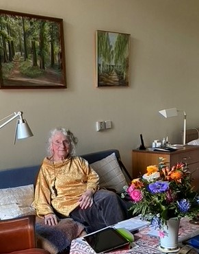 Eine Frau, Helga Bartsch, sitz auf einem blauen Sofa, das vor einer pastellgelben Wand steht. Vor ihr auf einem kleinen Tisch stehen eine Vase mit einem bunten Blumenstrauß.